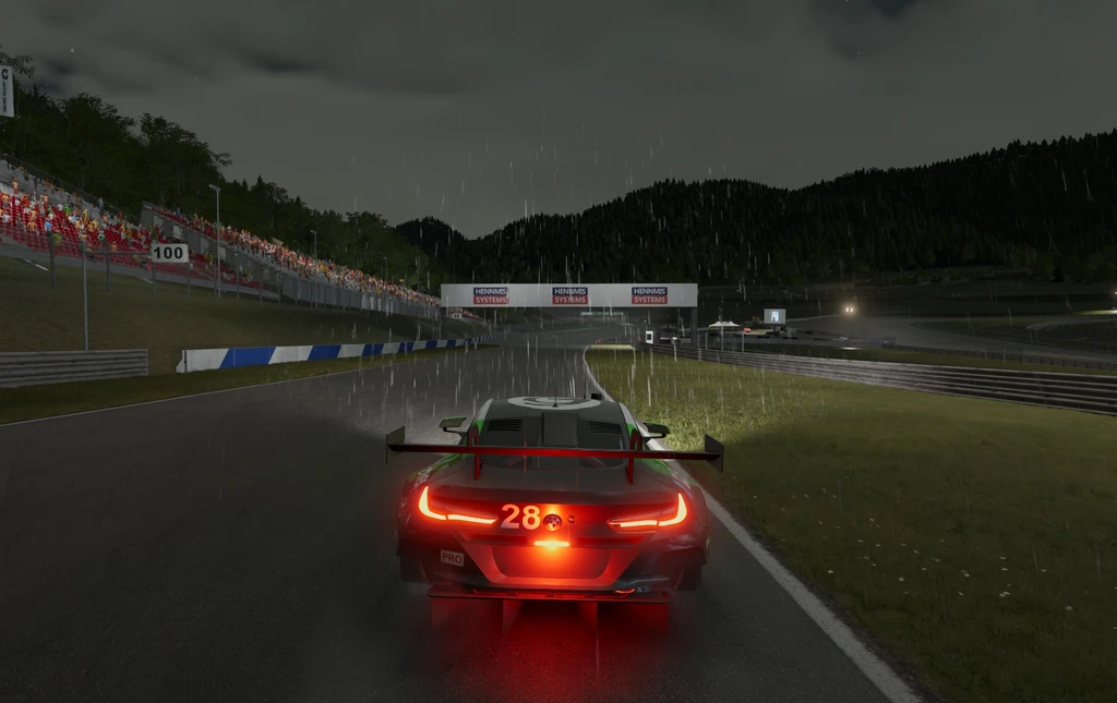Racerbil med nummer 28 och lysande bromsljus kör på en våt bana i regn och skymning, med läktare och skogsklädda berg i bakgrunden i Project Motor Racing.