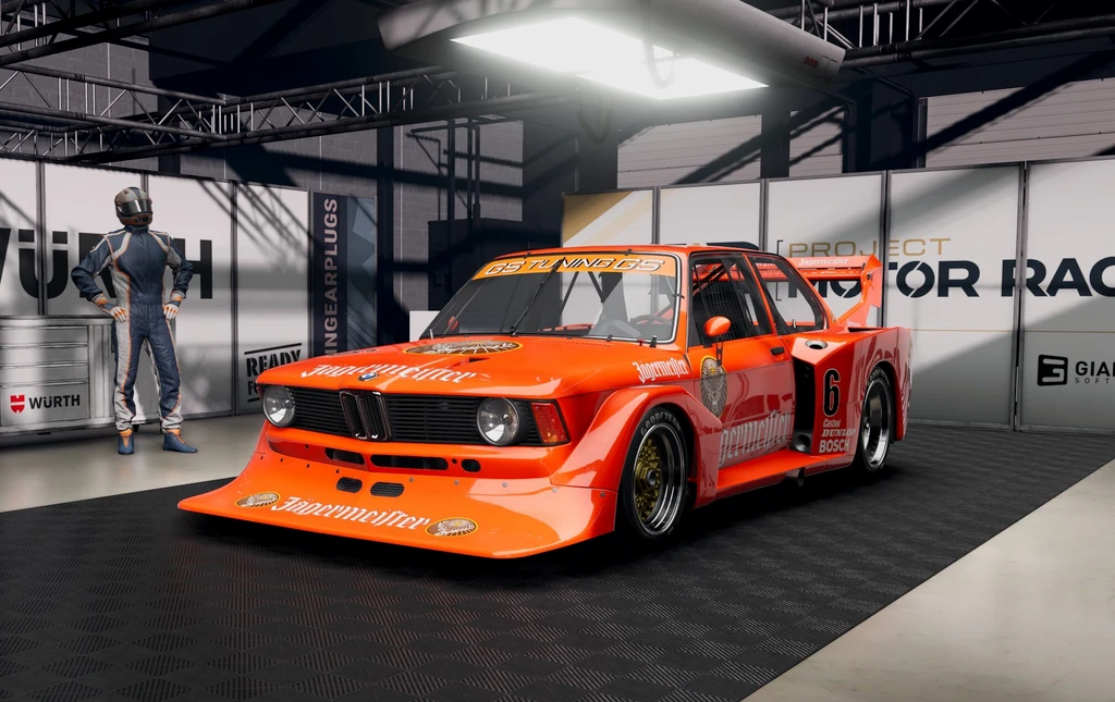 Orange BMW-racerbil med Jägermeister-dekor och bred kaross står i ett garage i Project Motor Racing, med en förare i overall bredvid.