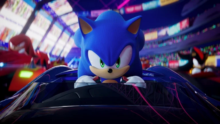 Sonic the Hedgehog sitter i sin blå racerbil med blicken fäst framåt, i en upplyst arena med publik. Knuckles och Shadow syns suddigt i bakgrunden.