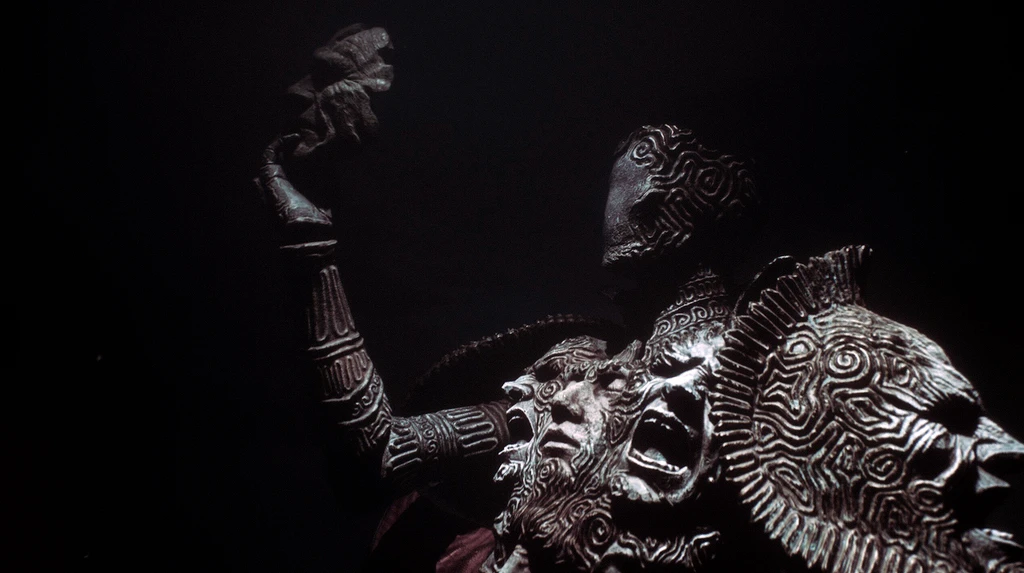 Mörk scen med en ornamenterad bronsliknande staty av en gestalt med mönstrad mask som höjer en hand, framför skrikande ansikten och en spiralmönstrad sköld.