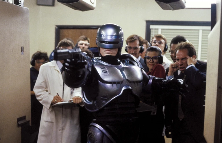 Robocop i silvergrå rustning sträcker fram pistolen på en skjutbana, omgiven av hörlursförsedda åskådare i kostym och labbrock, varav en håller för örat.
