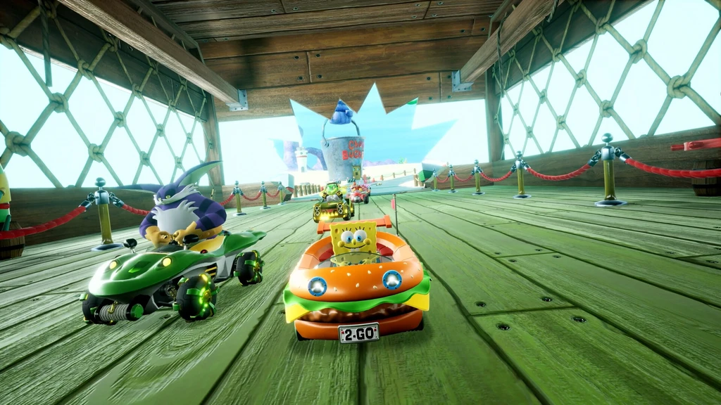 Svampbob i en hamburgarformad kart och Big the Cat i en grön kart tävlar sida vid sida över ett trädäck med rep och räcken i Sonic Racing: CrossWorlds.