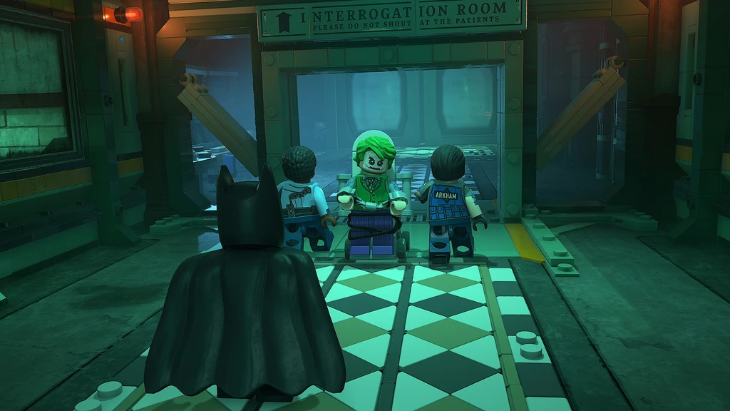 Lego Batman: Legacy of the Dark Knight: Batman står med ryggen mot oss i ett grönskimrande rum och tittar på Jokern, som sitter i rullstol mellan två Arkham-vakter under skylten Interrogation Room.