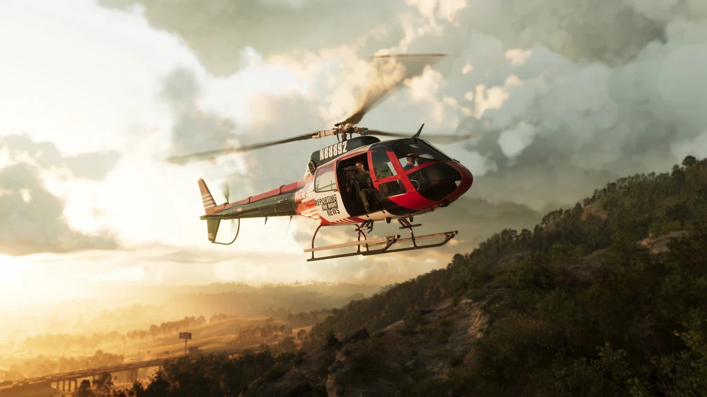 Röd och vit helikopter med öppen sidodörr flyger över en skogsklädd bergssluttning i gyllene motljus, med dramatiska moln och en motorvägsbro i dalen nedanför.