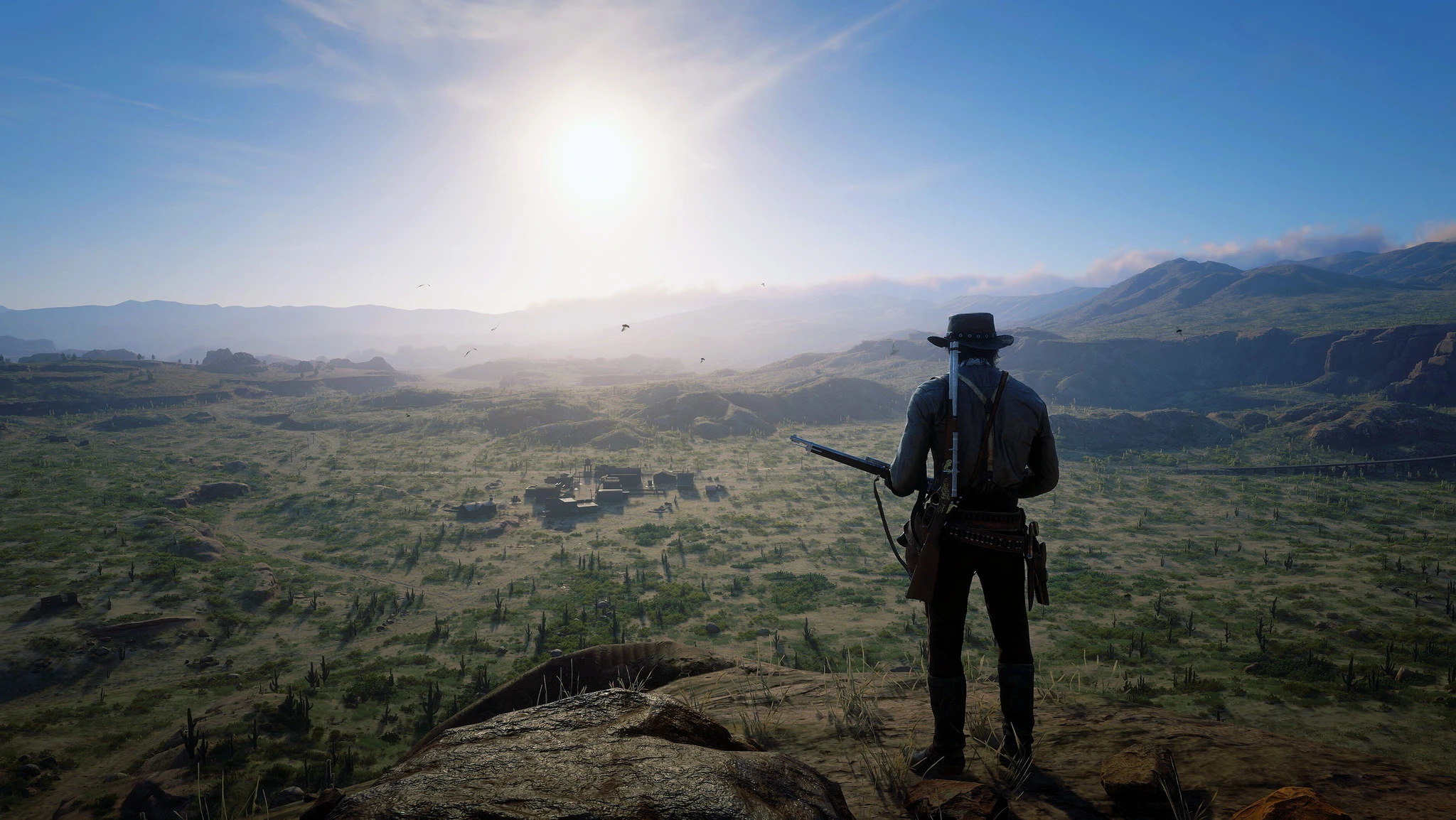 Cowboy med gevär i handen står på en klippa i Red Dead Redemption 2 och blickar ut över en solbelyst dal med en liten by i fjärran.