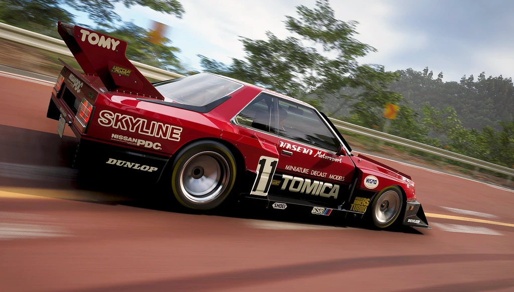 Röd Nissan Skyline i racingutförande med Tomica- och Tomy-dekaler tar en kurva i hög fart på en landsvägsbana omgiven av träd.
