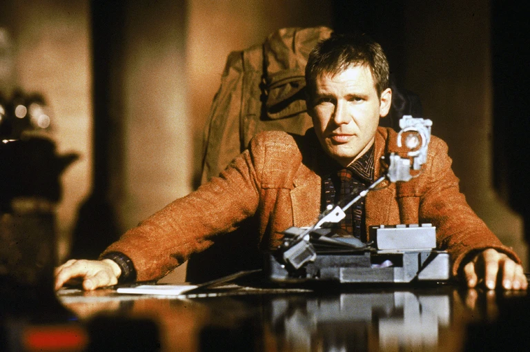 Harrison Ford sitter vid ett bord i en brun tweedjacka och ser rakt in i kameran, framför en apparat med liten rörlig arm. Scen ur Blade Runner i dunkelt, varmt ljus.