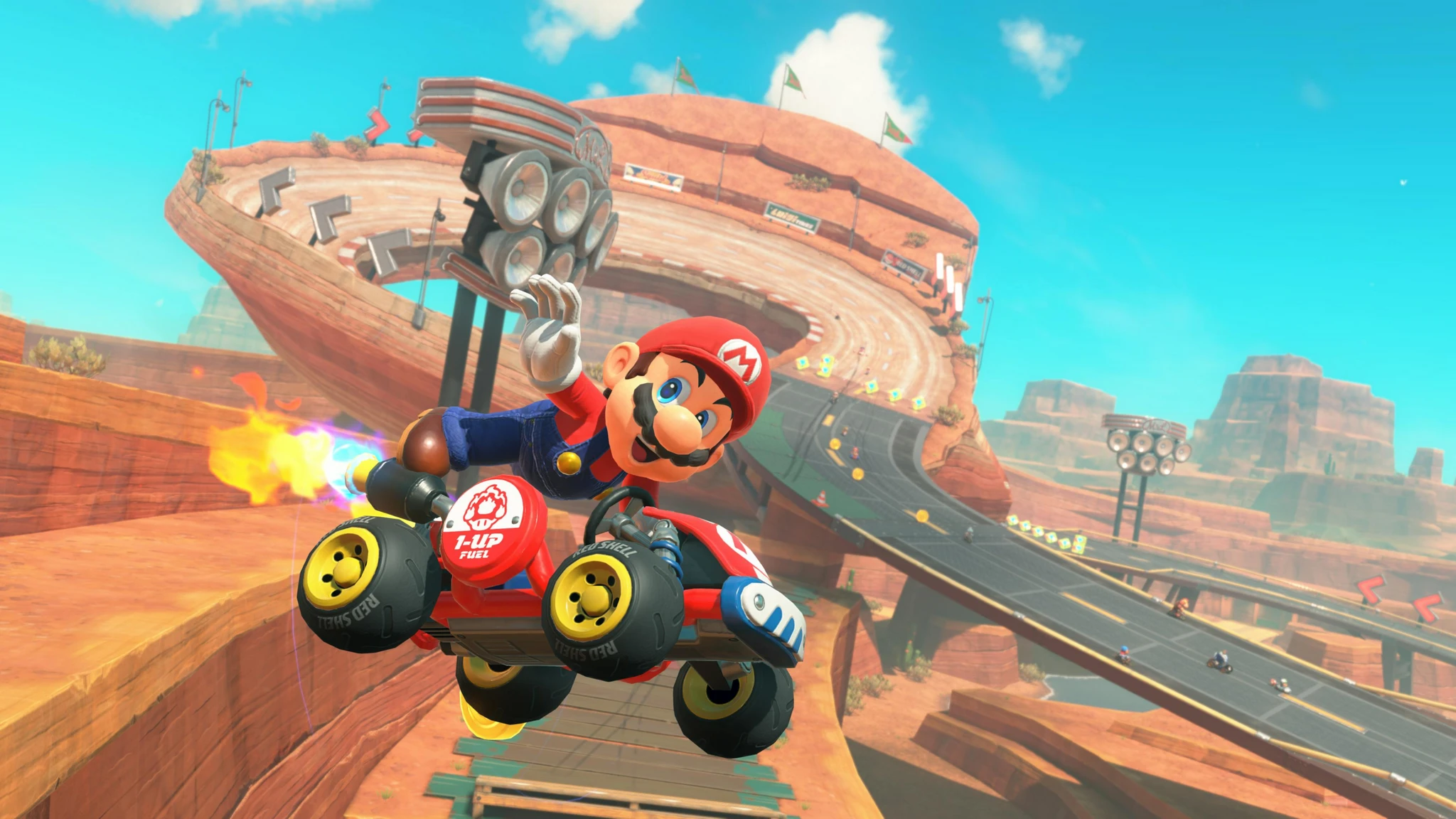 Mario i sin röda kart hoppar högt över en bana i en röd ökenmiljö, med en slingrande väg och klippformationer i bakgrunden och en eldflamma bakom karten.