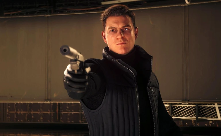 En allvarlig man i svart väst och handskar riktar en pistol med ljuddämpare rakt mot kameran i en industriell hall, en scen ur 007 First Light.