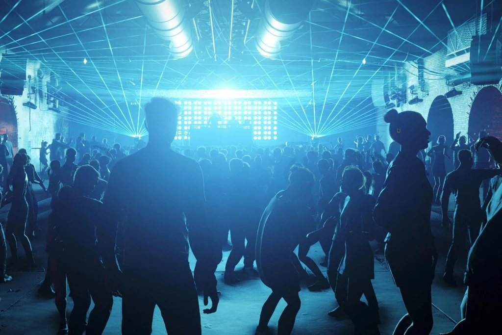 Silhuetter av dansande folkmassa i en stor klubblokal, med blå laserstrålar och ett ljusbås med DJ i bakgrunden.