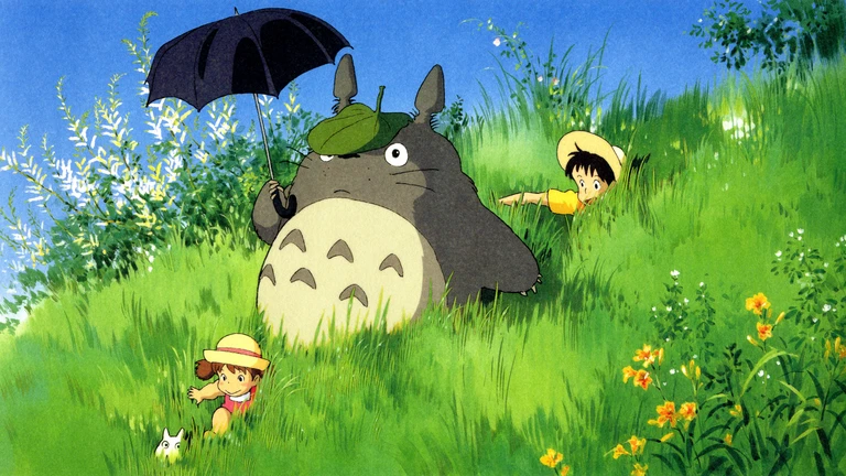 Totoro med ett löv på huvudet och ett svart paraply står på en grön, blommande äng medan två barn i strålhattar kryper i gräset runt honom.