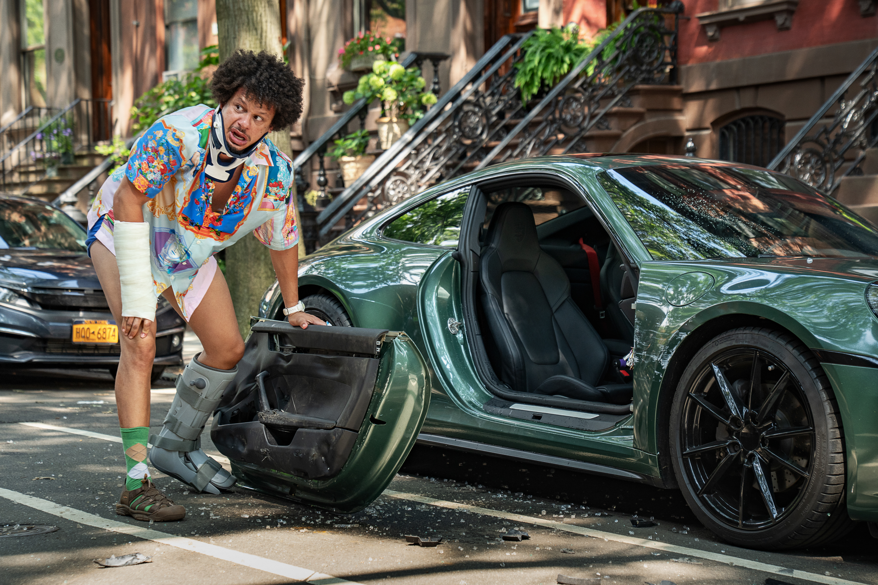 "Injured man in colorful clothes stands on a city street next to a green sports car with the driver’s door ripped off, broken glass on the ground, and brownstone buildings in the background."