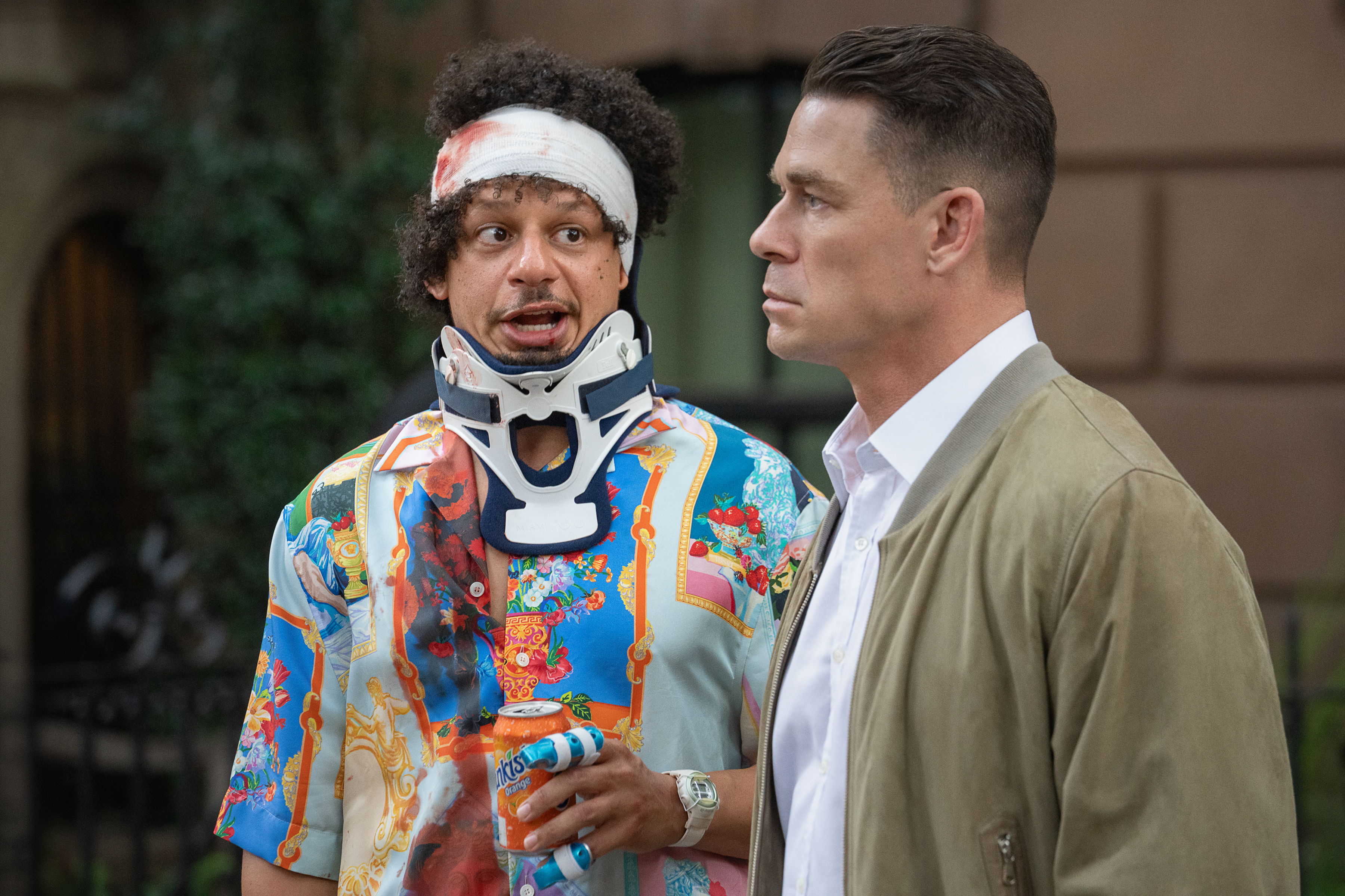 "Two men outdoors: one wears a colorful shirt, head bandage, and neck brace, holding a can, looking surprised; the other in a tan jacket and white shirt, looking serious. Urban background with greenery and brownstone building."