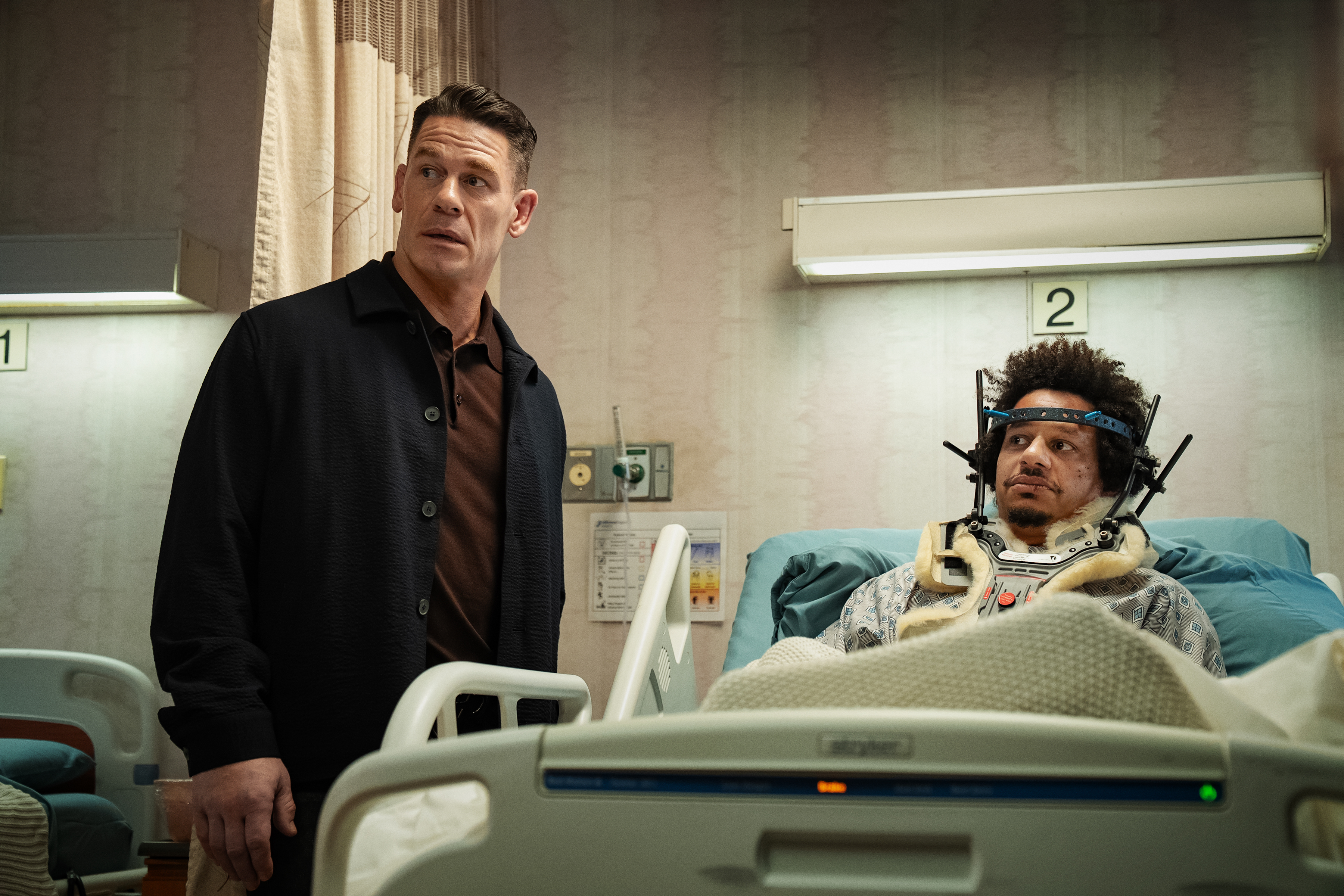 "A man stands beside a hospital bed looking surprised while another person lies in bed with medical headgear, in a hospital room setting with numbered beds and medical equipment visible."