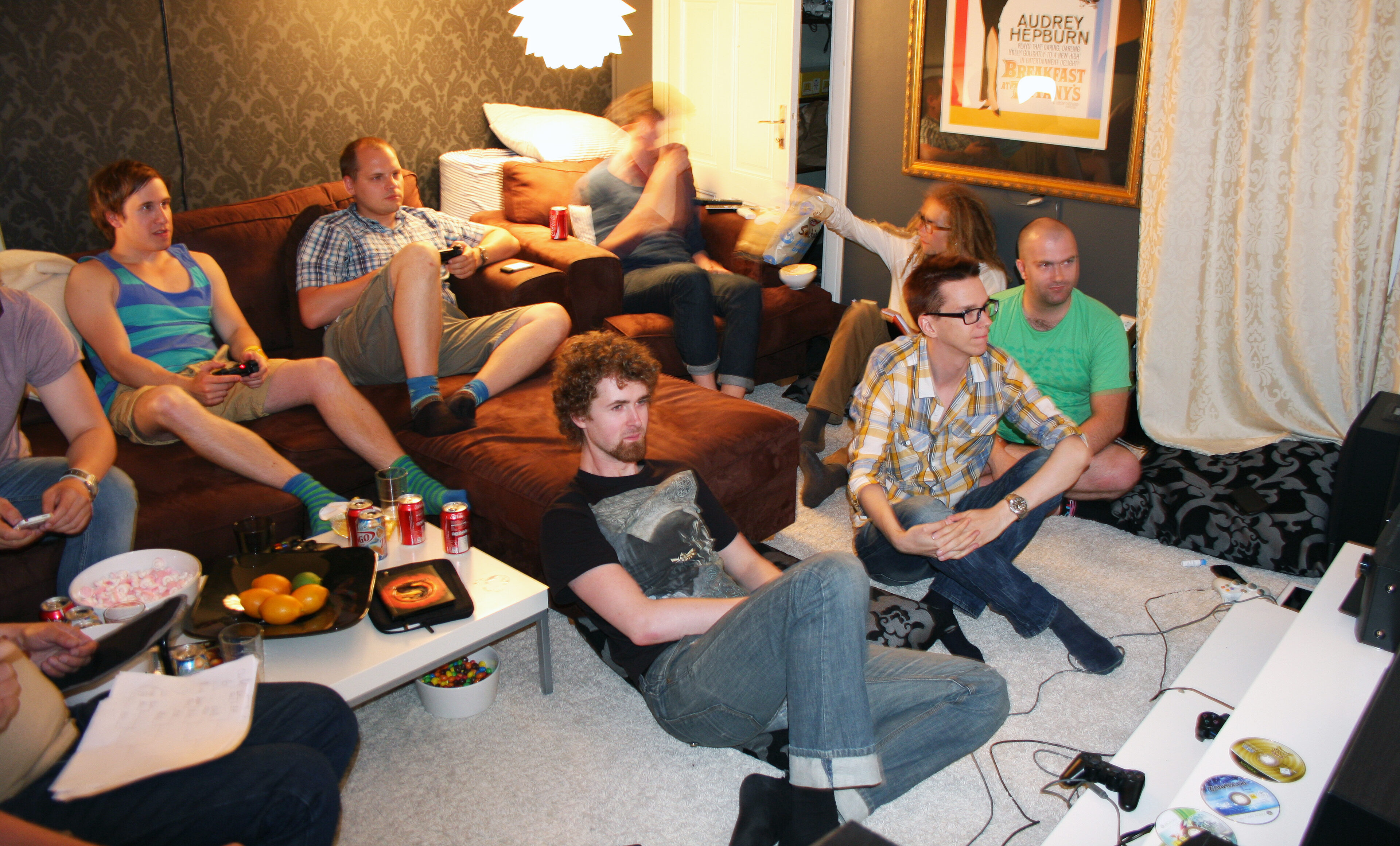 Åtta vänner och kollegor samlade i en vardagsrum för spelafton, sittande i brun soffa och på golvet med läsk och snacks på soffbordet framför en tv.