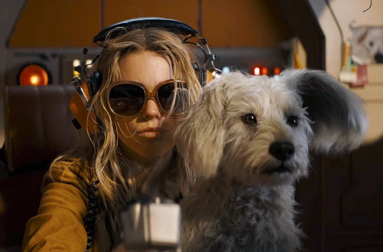 Ung kvinna med rufsigt blont hår, hörlurar och stora solglasögon sitter i en trång cockpit med en lurvig grå hund tätt intill sig.