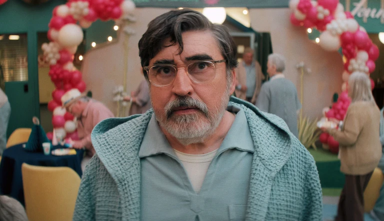 Sam, spelad av Alfred Molina, står med allvarlig min i ljusblå kofta och glasögon framför ballongbågar i rött och vitt, med äldre personer i bakgrunden, i serien The Boroughs.