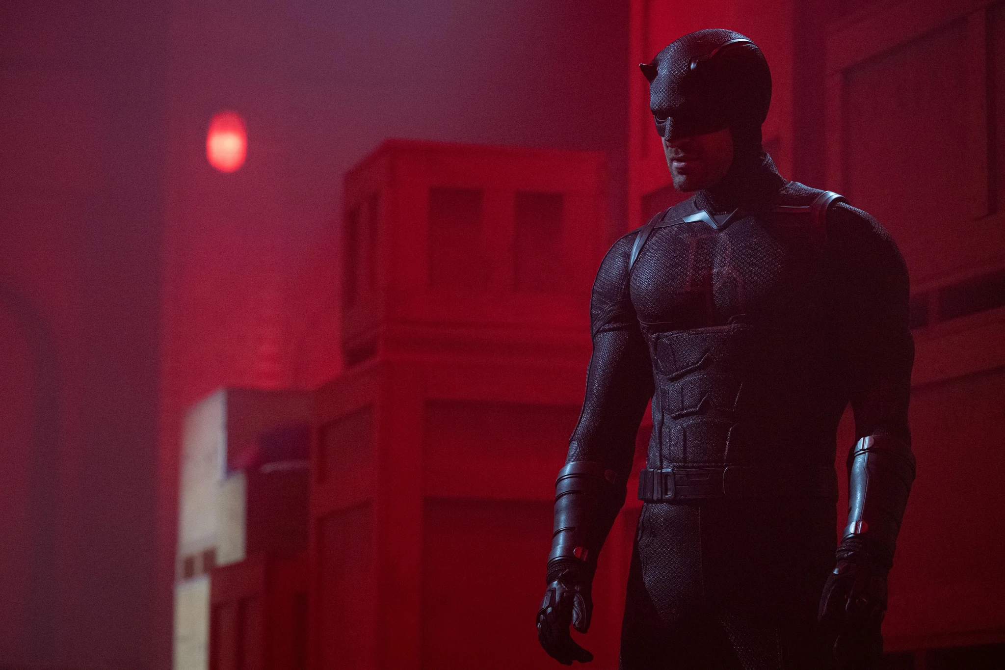 Daredevil i mörk dräkt och hjälm med horn står på ett tak i rödbelyst dis, med takuppbyggnader i bakgrunden, i Daredevil: Born Again.