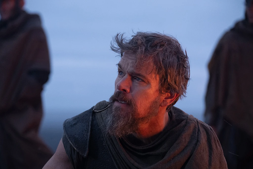 Skäggig man med rufsigt hår och grov mantel tittar åt sidan i skymningsljus, med oskarpa kappklädda gestalter bakom sig, i en scen från The Odyssey.