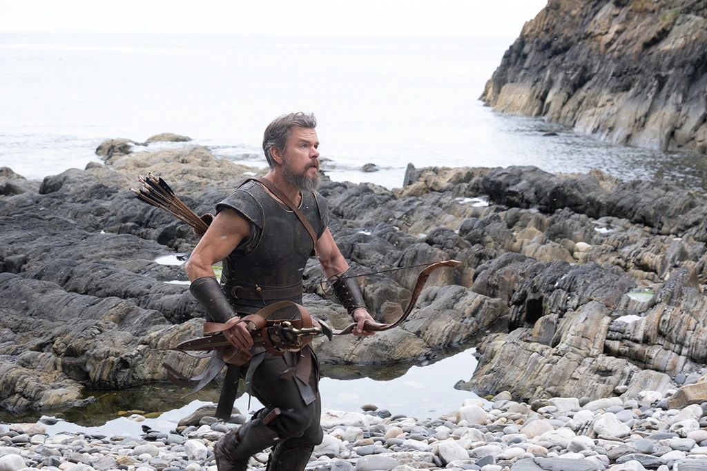 Skäggig man i läderrustning med pilkö på ryggen går över klippor vid en stenstrand och håller en stor båge i handen, i en scen från The Odyssey.