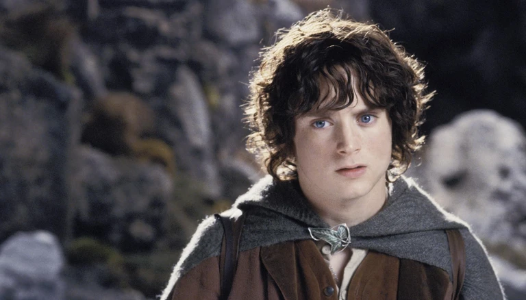 Närbild på en ung man med mörkt, lockigt hår och blå ögon, klädd i grå huva med bladformad spänne och brun väst, framför en stenig bergsvägg i The Lord of the Rings: The Two Towers.