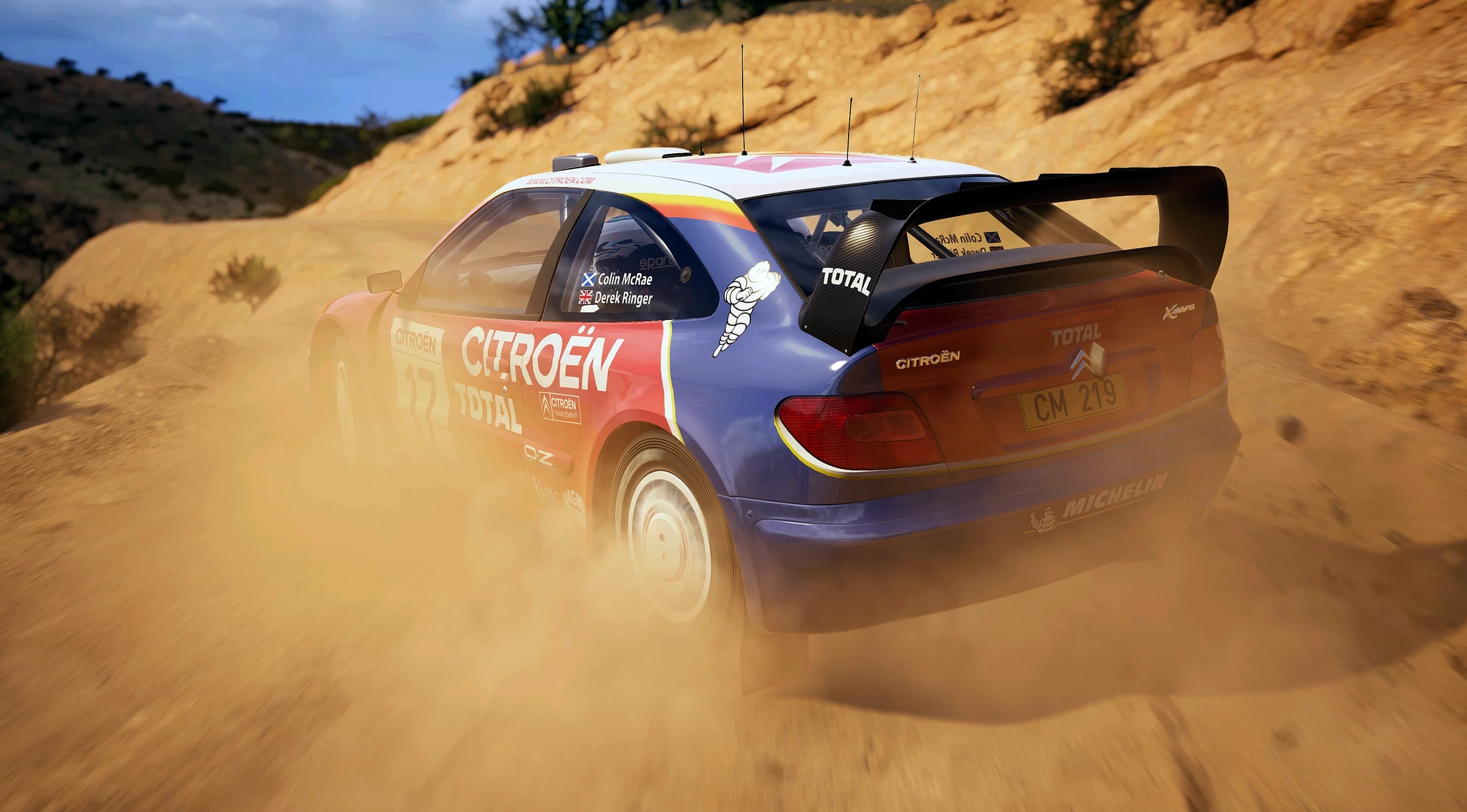 Blå och röd Citroën rallybil med stor bakvinge driftar genom en dammig grusväg i en torr, kuperad miljö i EA Sports WRC.