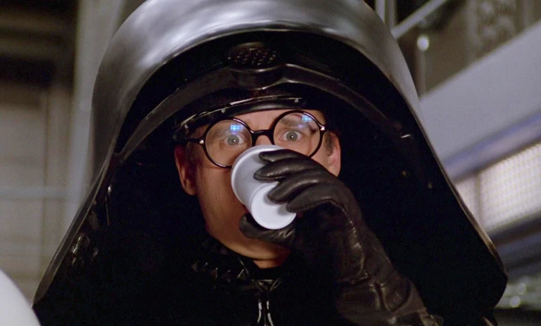 Man i en enorm svart hjälm och rundade glasögon dricker ur en vit plastmugg med storögd blick, en scen ur Spaceballs.
