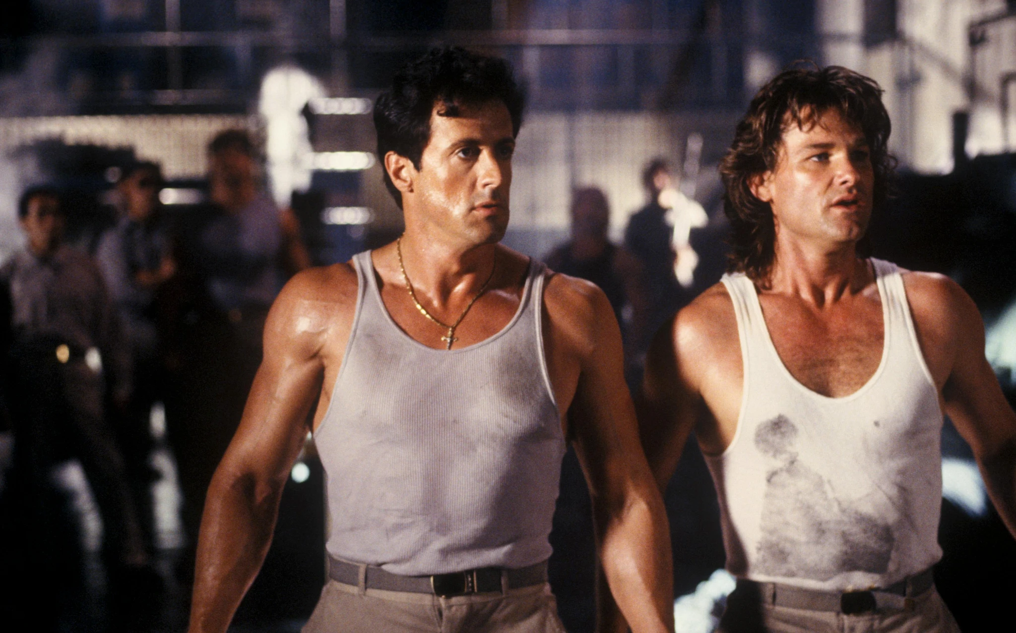 Två svettiga, muskulösa män i linnen står sida vid sida i en dunkel lokal med suddiga personer i bakgrunden, en mörkhårig med guldkedja och en långhårig i vitt linne. Scen ur Tango & Cash.