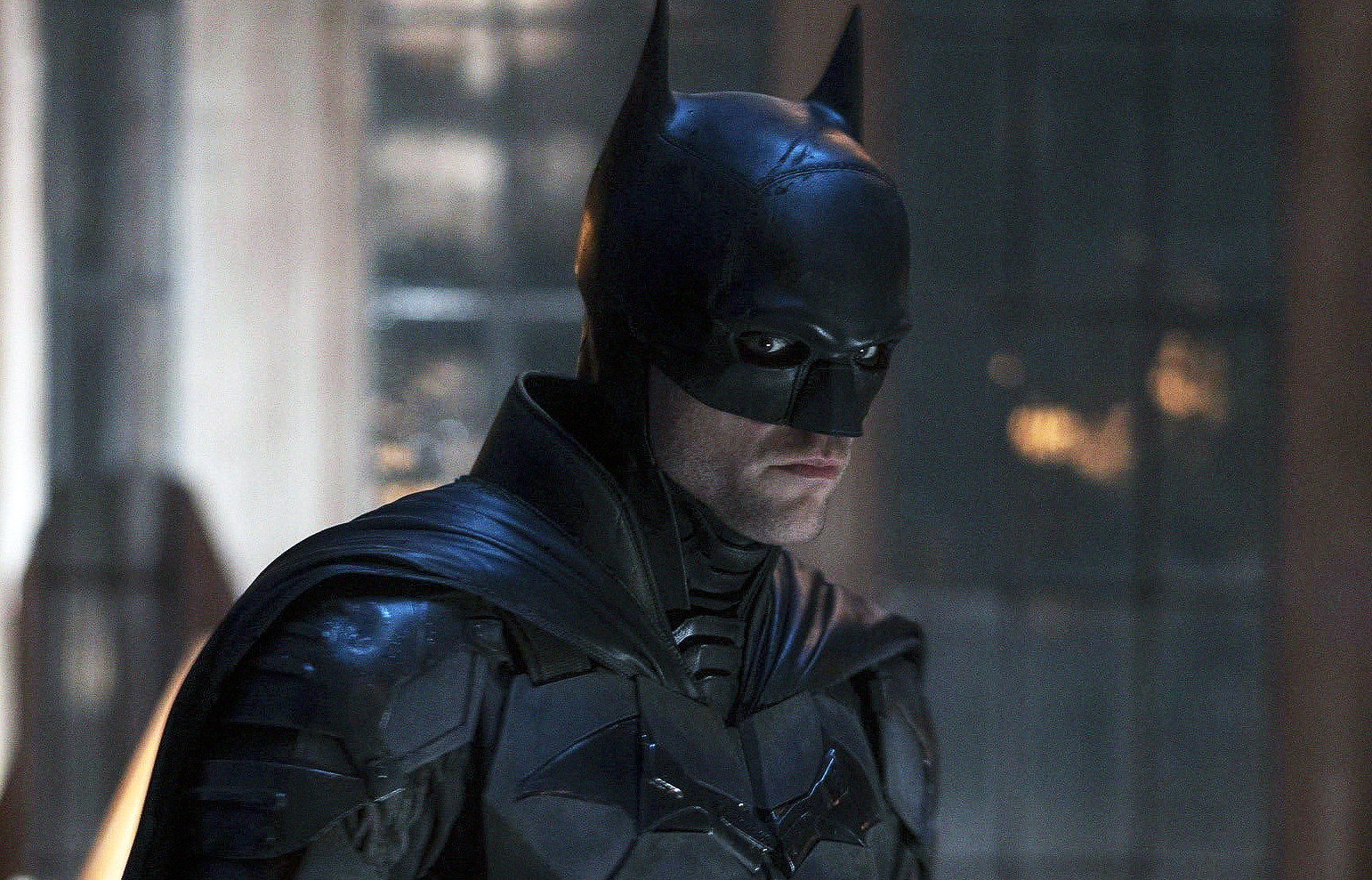 Batman i svart dräkt med spetsig mask och mantel blickar allvarligt åt sidan i ett dunkelt, regnigt rum med suddig bakgrund.