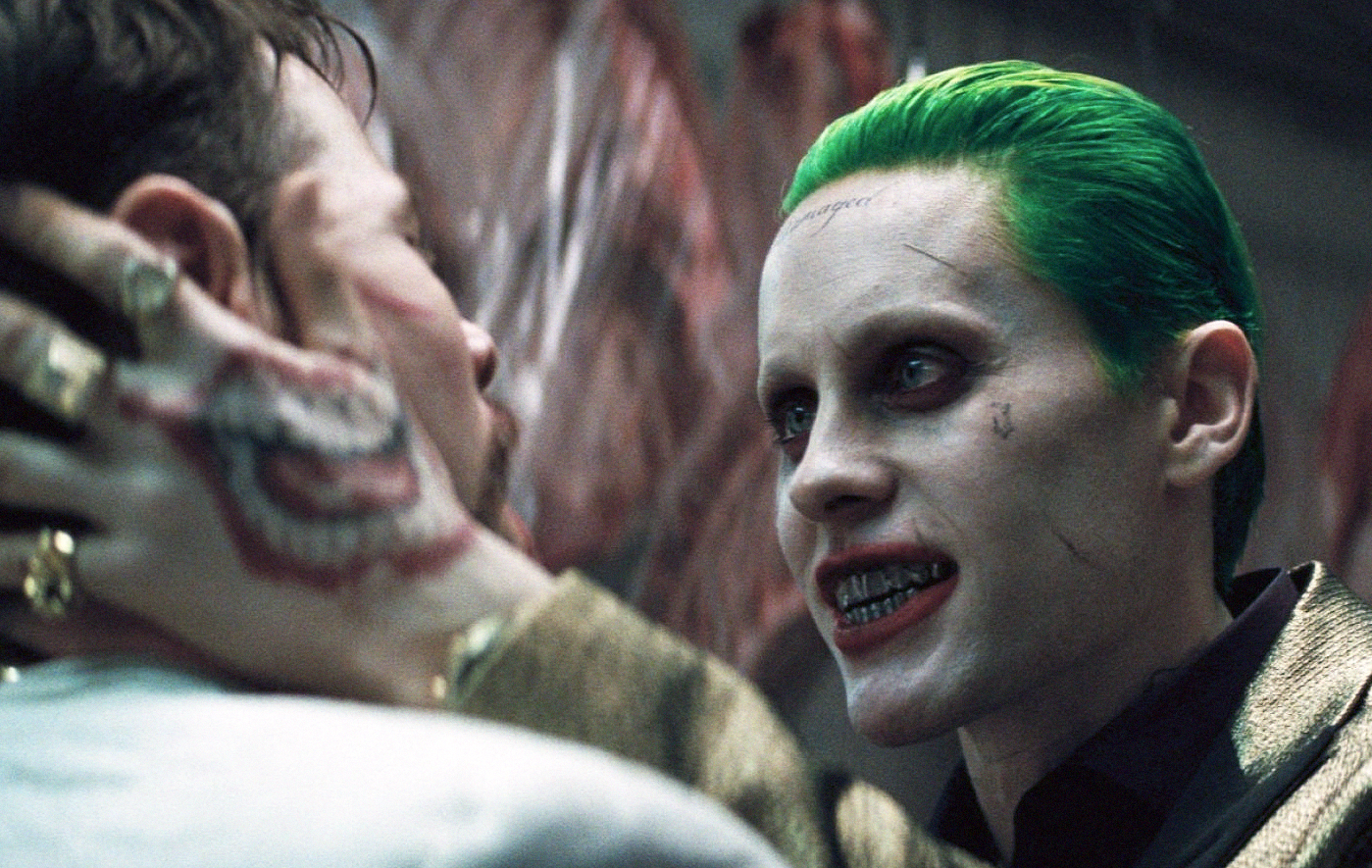 Joker med grönt hår, vitsminkat ansikte och metalltänder håller en man hårt i ansiktet med en tatuerad hand och stirrar honom nära in på livet.