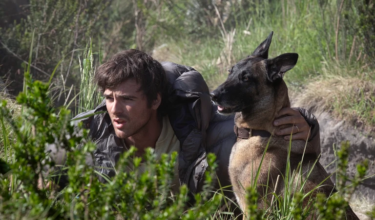 En mörkhårig man i läderjacka kryper ihop i högt gräs och buskar med en schäferhund vid sin sida. Båda spanar spänt åt samma håll i filmen The Dog Stars.