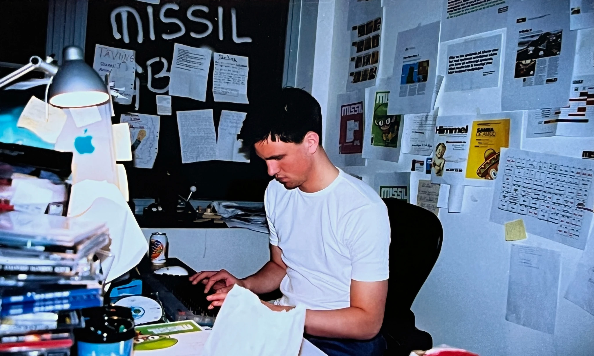 Ung man i vit t-shirt sitter vid ett skrivbord fyllt av spel-cd och papper och skriver på tangentbordet. På väggarna hänger utklipp och uppslag, och på tavlan står "Missil".