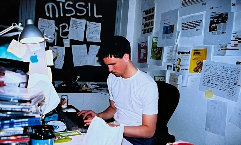 Ung man i vit t-shirt sitter vid ett skrivbord fyllt av spel-cd och papper och skriver på tangentbordet. På väggarna hänger utklipp och uppslag, och på tavlan står "Missil".