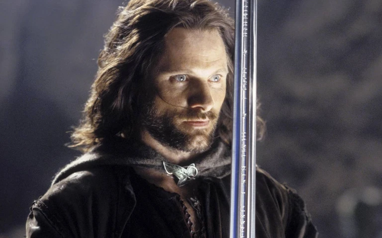 Man med långt mörkt hår och skägg, klädd i mörk mantel med spänne, blickar allvarligt åt sidan bakom ett lodrätt svärd med inristade tecken i The Lord of the Rings: Return of the King.