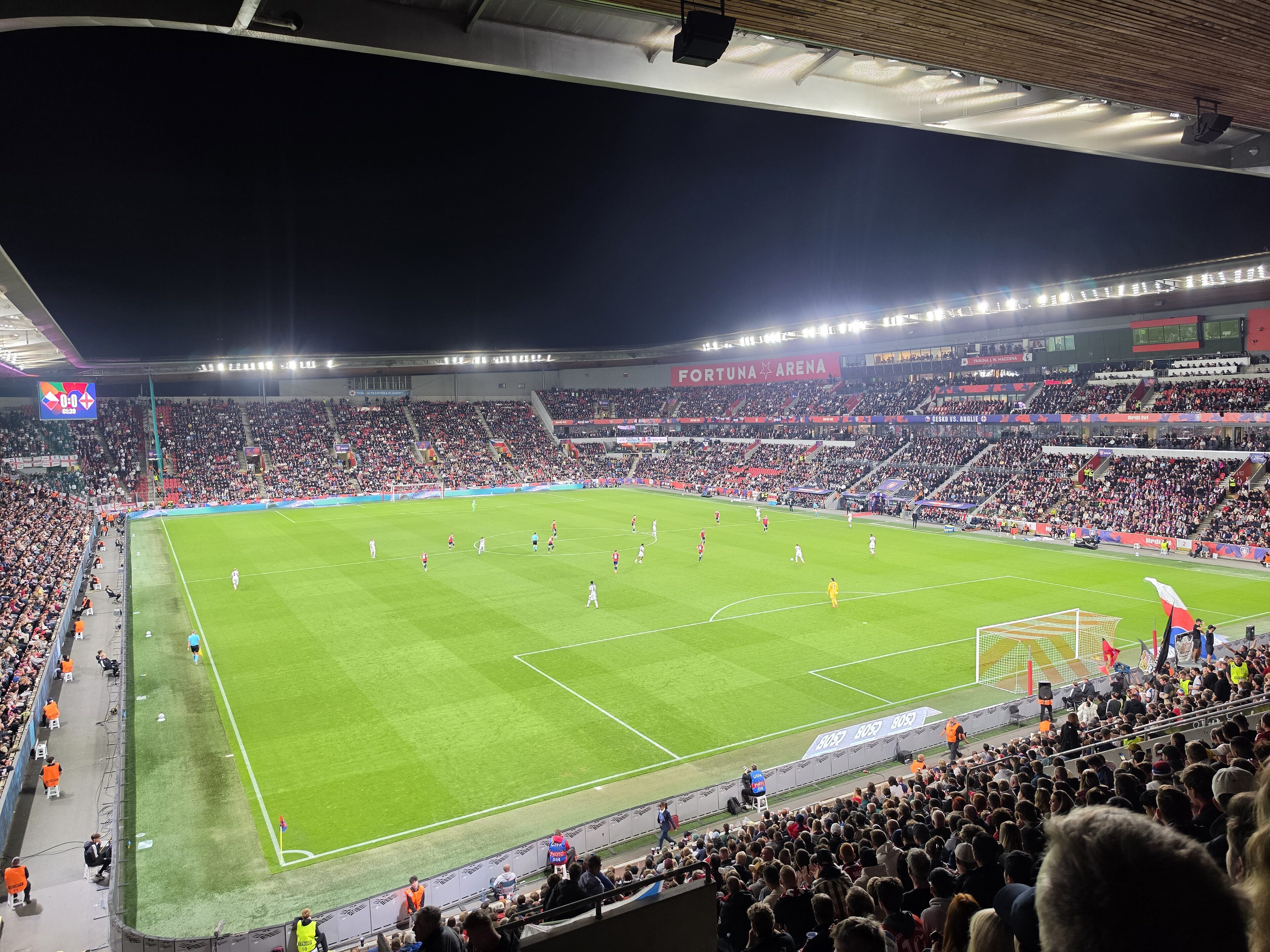 Fotbollsmatch på en upplyst arena på kvällen med fullsatta läktare och spelare i röda och vita dräkter på en grön gräsplan.