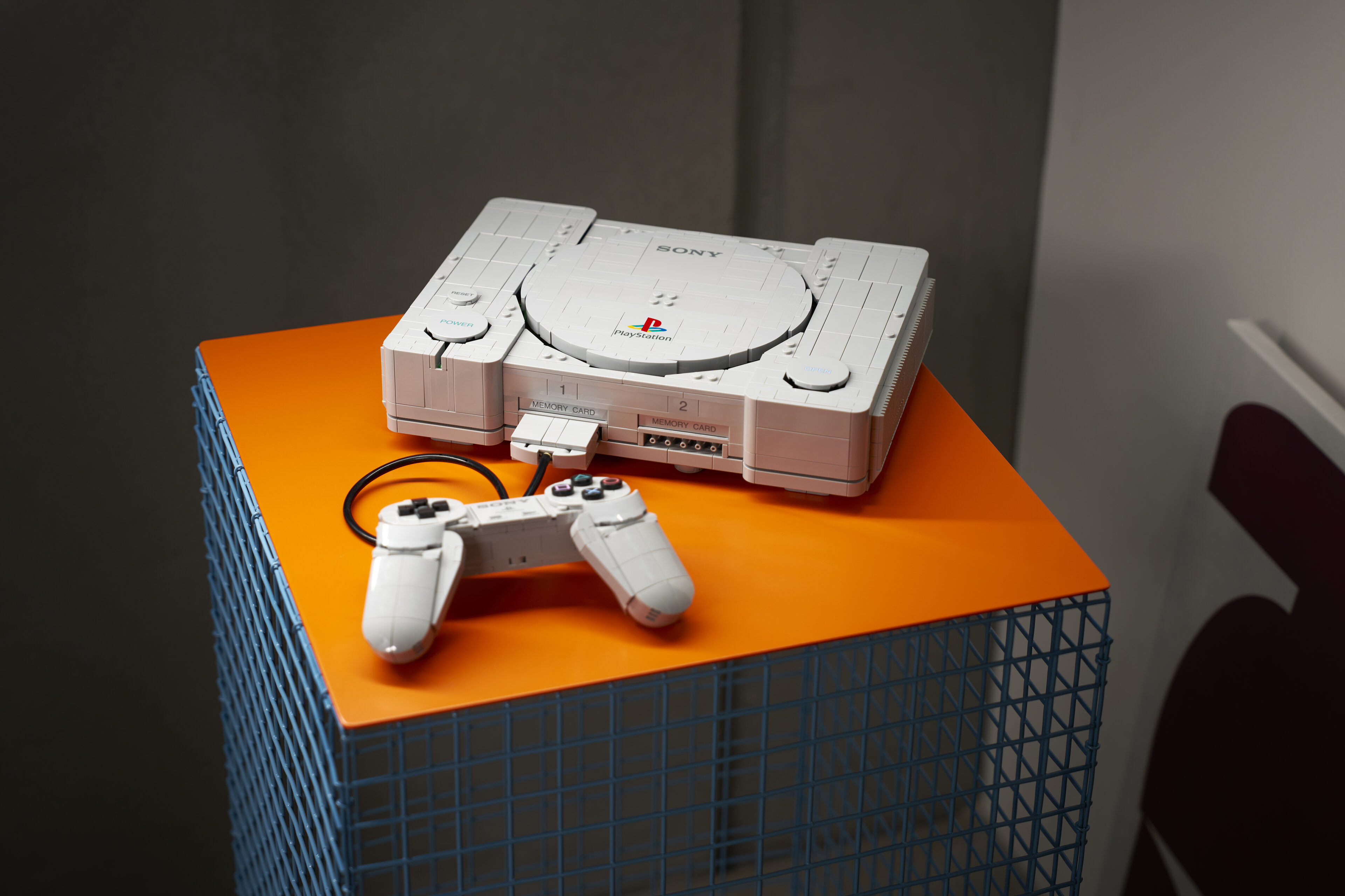 Lego-modell av den första Playstation-konsolen med tillhörande handkontroll, placerad på ett orange podium med blått ställ av metallgaller.