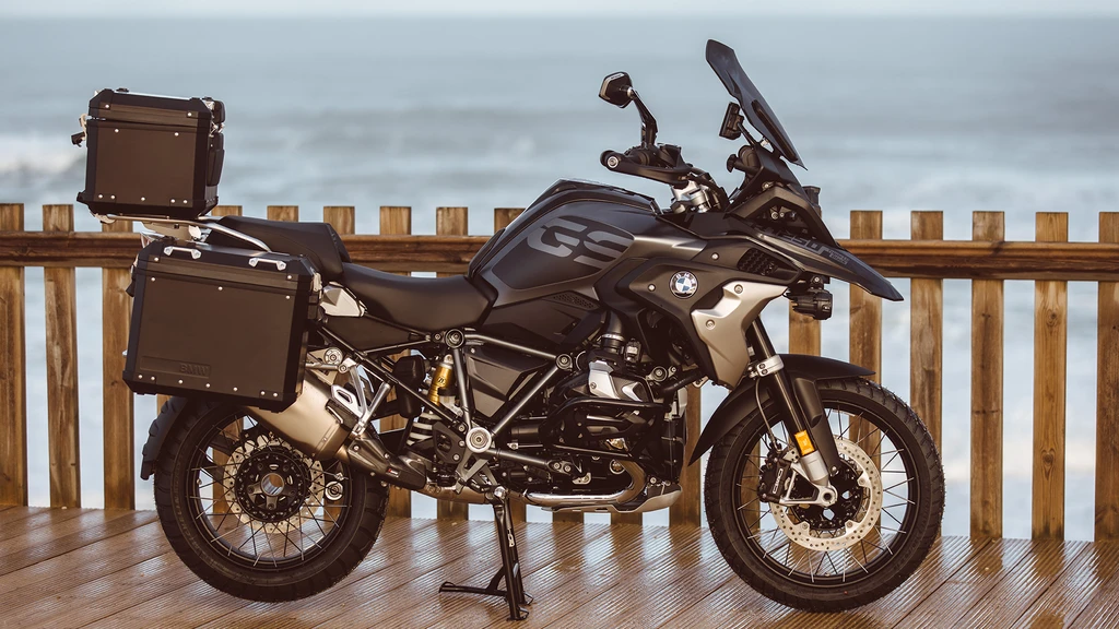 Svart BMW GS-äventyrsmotorcykel med sidokoffertar och toppbox, uppställd på ett vått trädäck framför ett staket och havet.