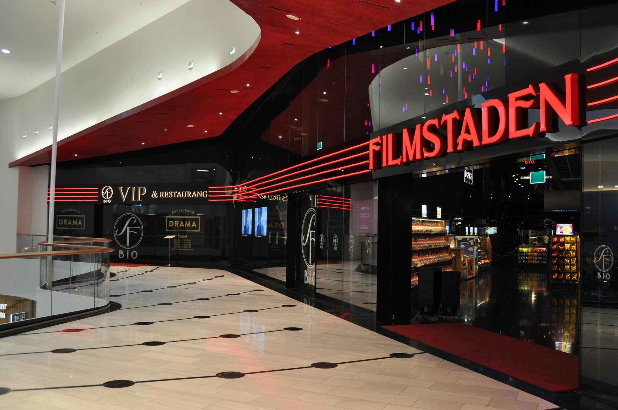 Entrén till Filmstaden med röd neonskylt, mörk glasfasad och en VIP-avdelning med restaurang, sedd från ett ljust köpcentrum med blankt golv.