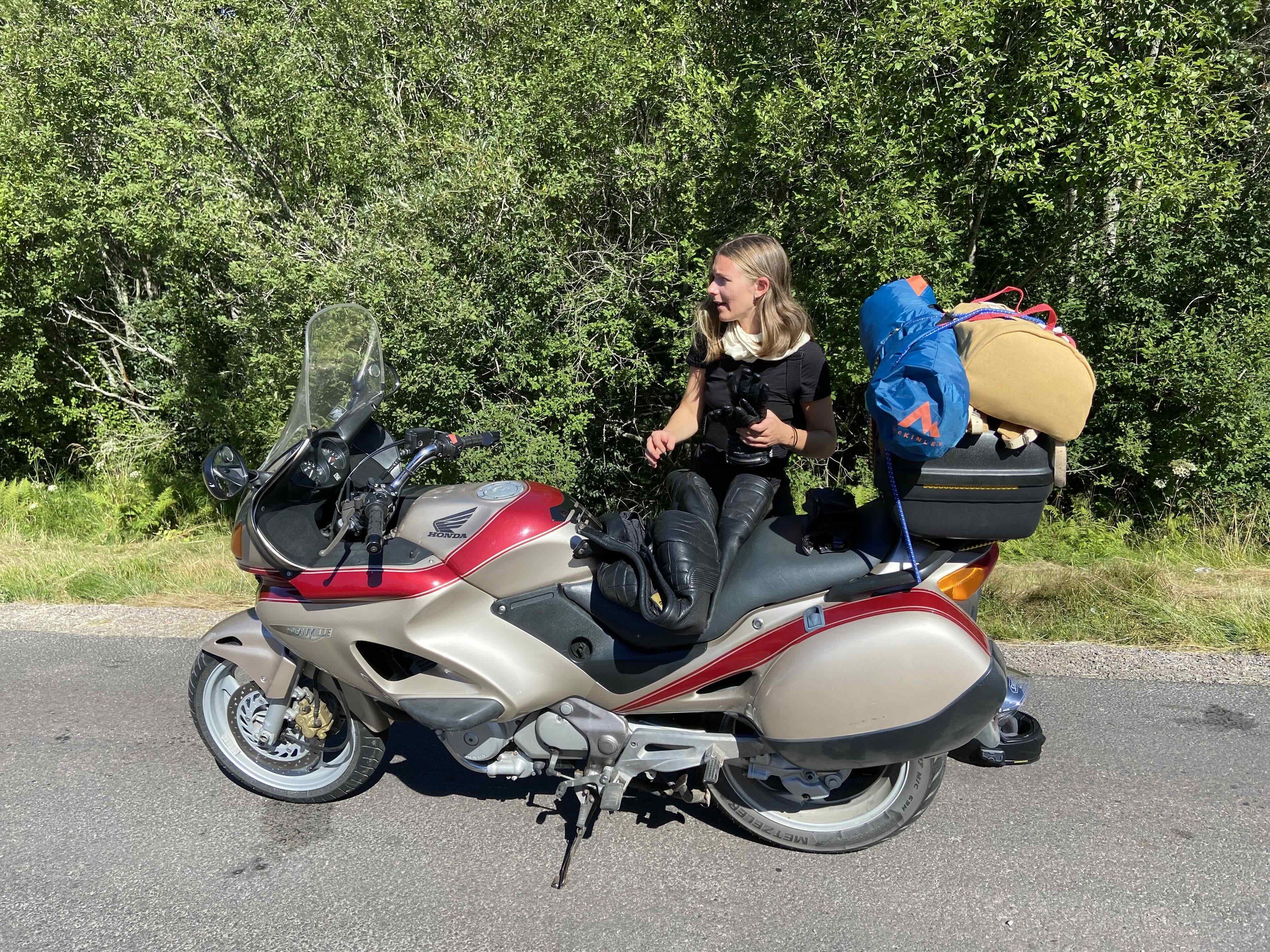 Amanda står bakom en beige och röd Honda Deauville från 1998 på en landsväg, med packning och ett blått tält högt uppe på pakethållaren.