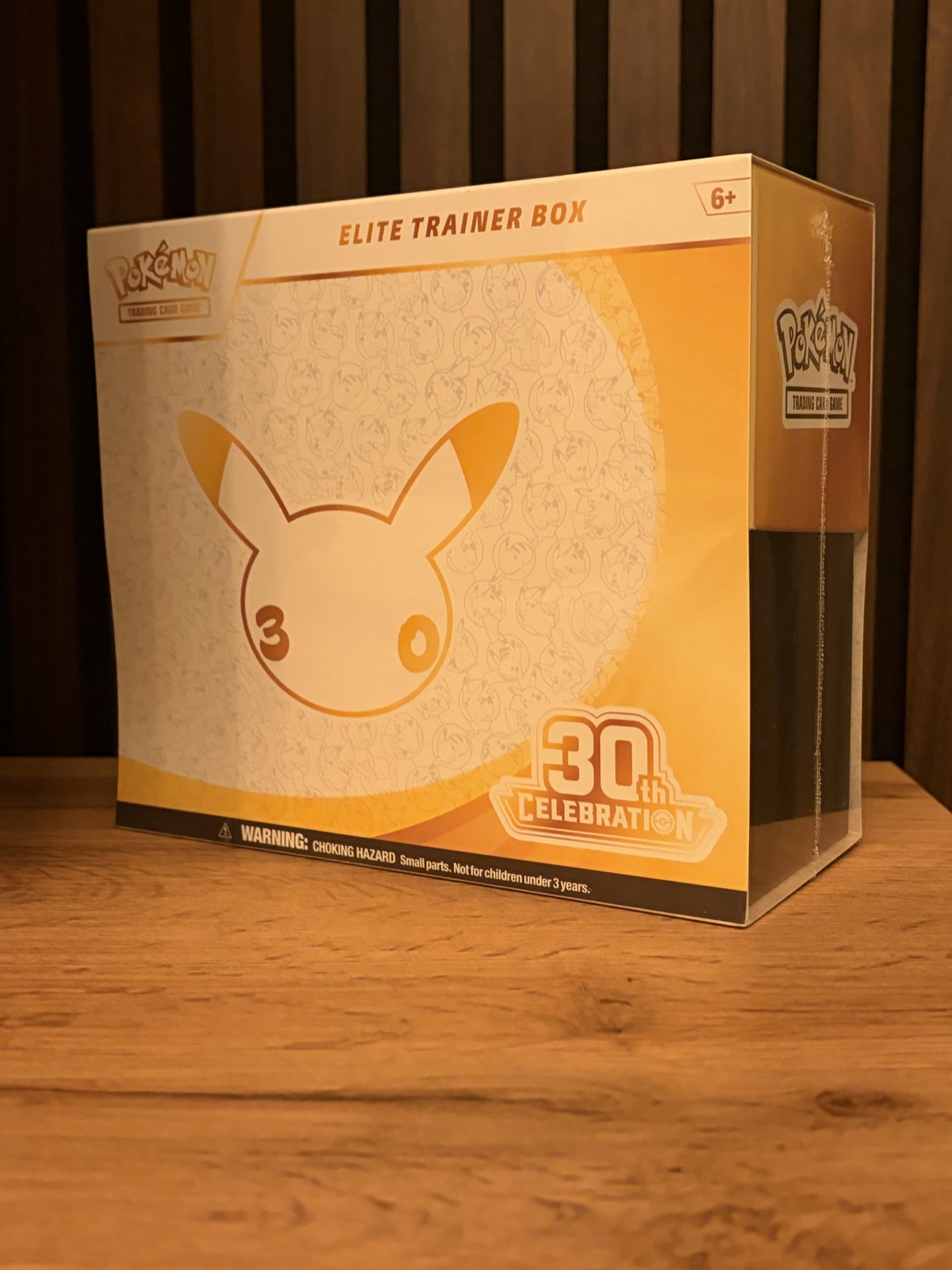 Pokémon Elite Trainer Box för 30-årsjubileet i guld och vitt, med en Pikachu-silhuett på framsidan, stående på ett träbord.