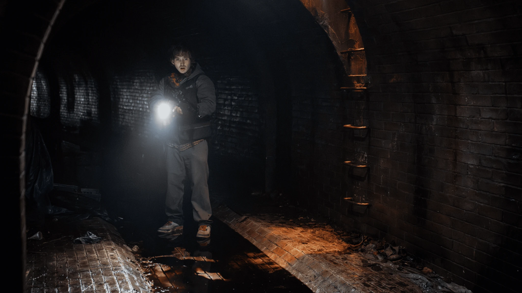 En ung man i huvtröja står i en mörk tegeltunnel och lyser med en ficklampa, med blicken spänt riktad framåt. Marken är blöt och skräpig.