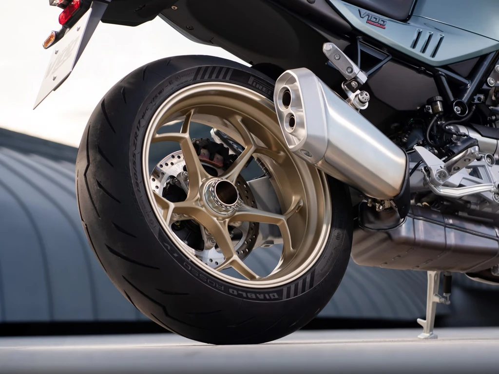 Närbild på bakhjulet på en Moto Guzzi V100 med guldfärgade fälgar, Pirelli Diablo-däck, silverfärgat ljuddämpare och sidostöd uppfällt mot en grå byggnad.