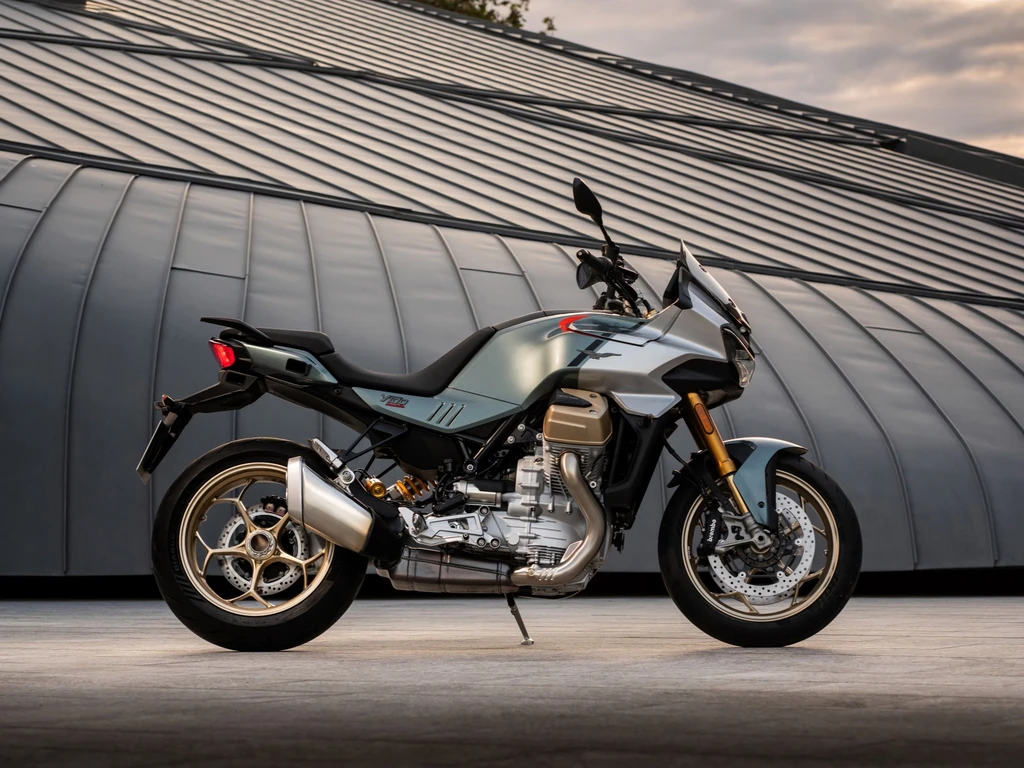 Silvergrå Moto Guzzi V100 sedd från sidan, med guldfärgade fälgar och synlig tvåcylindrig motor, parkerad framför en välvd metallbyggnad i skymning.