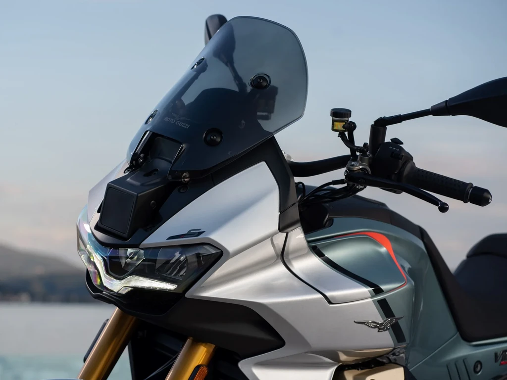 Närbild på framdelen av en silvergrå motorcykel med rökfärgat vindrutesglas, LED-strålkastare och guldfärgade framgafflar, med havet i bakgrunden.