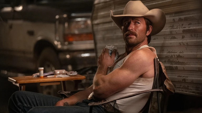 Man med mustasch och cowboyhatt, i linne och jeans, sitter i en fällstol framför en sliten husvagn och tittar bakåt över axeln. Bredvid står ett litet bord med medicinburk.