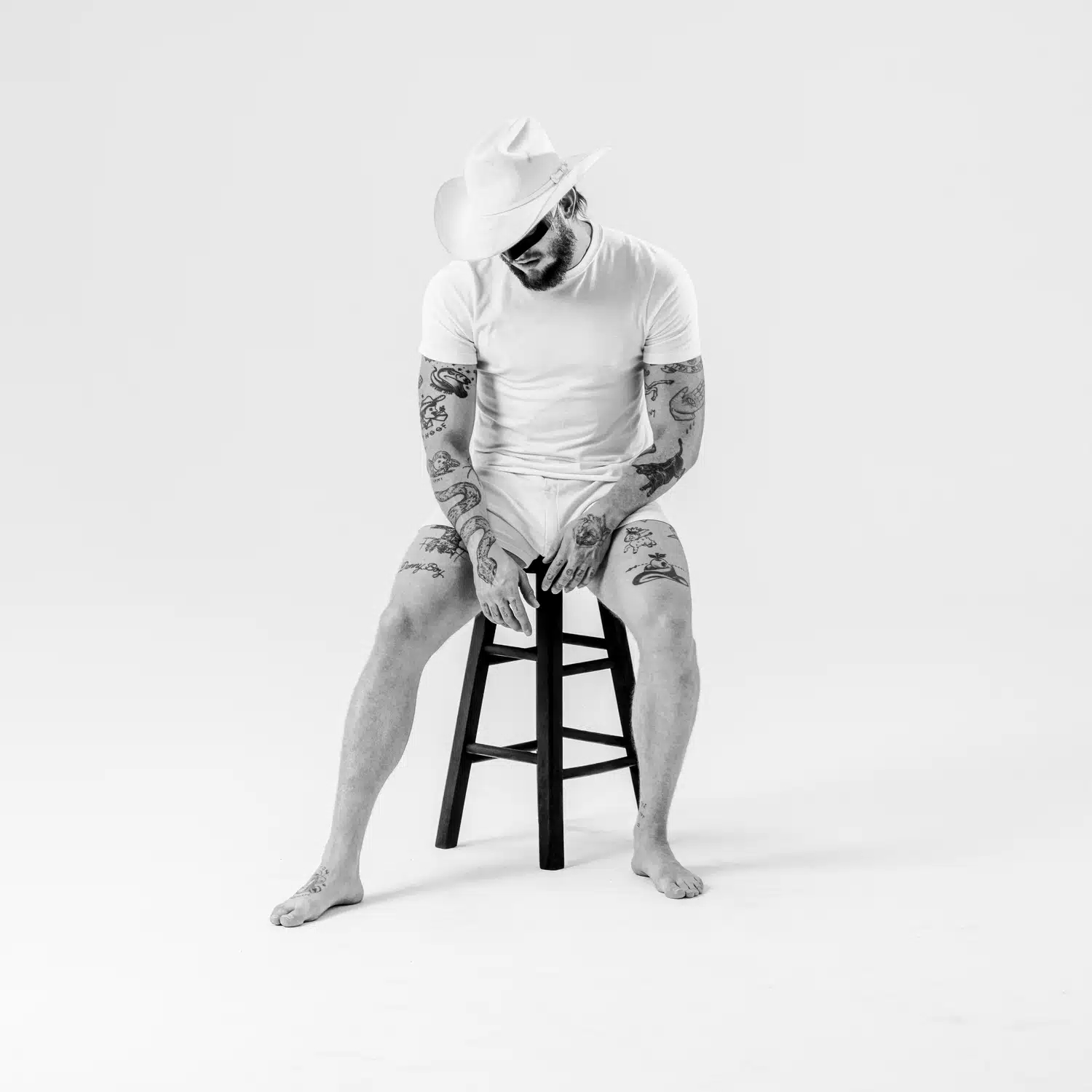 Orville Peck sitter barfota på en svart barstol i vit cowboyhatt och vit t-shirt, med tatuerade armar och ben och huvudet böjt framåt, mot vit bakgrund.