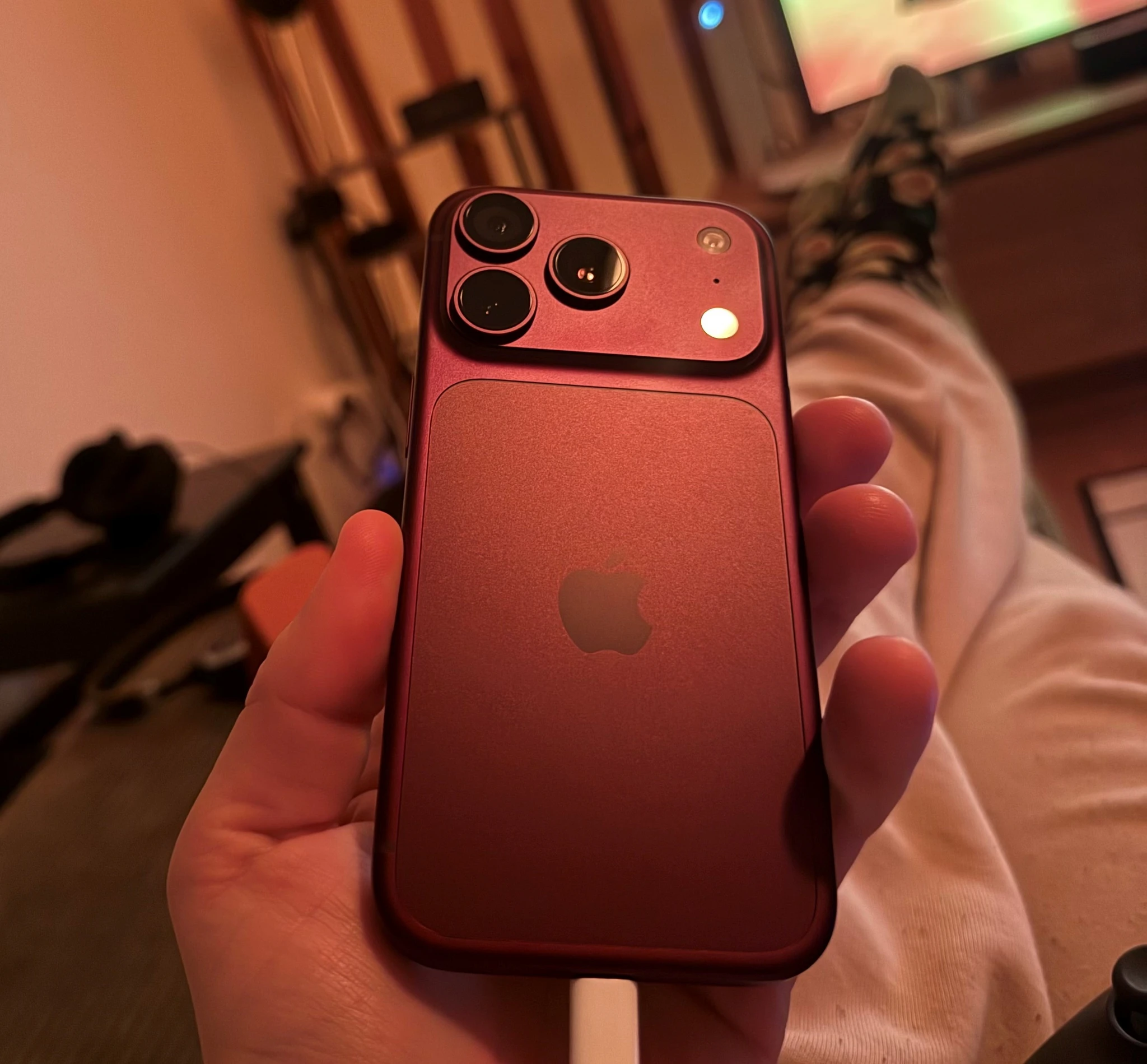 En hand håller upp en mörkröd iPhone med tre kameralinser och Apple-logotyp på baksidan, i ett dunkelt rum med en tv i bakgrunden.