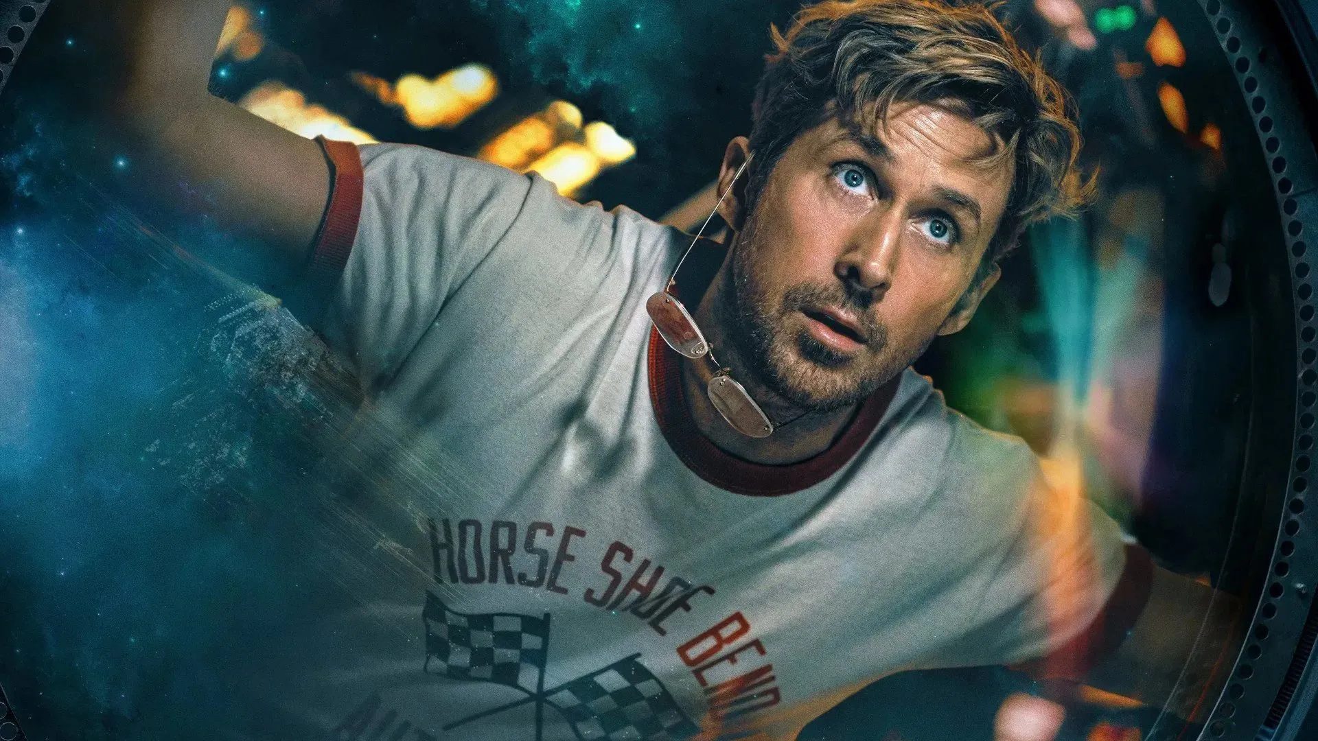 En skäggstubbig man med rufsigt blont hår och headset i en grå t-shirt svävar och tittar förundrat uppåt, omgiven av rymd och suddiga färgade ljus.