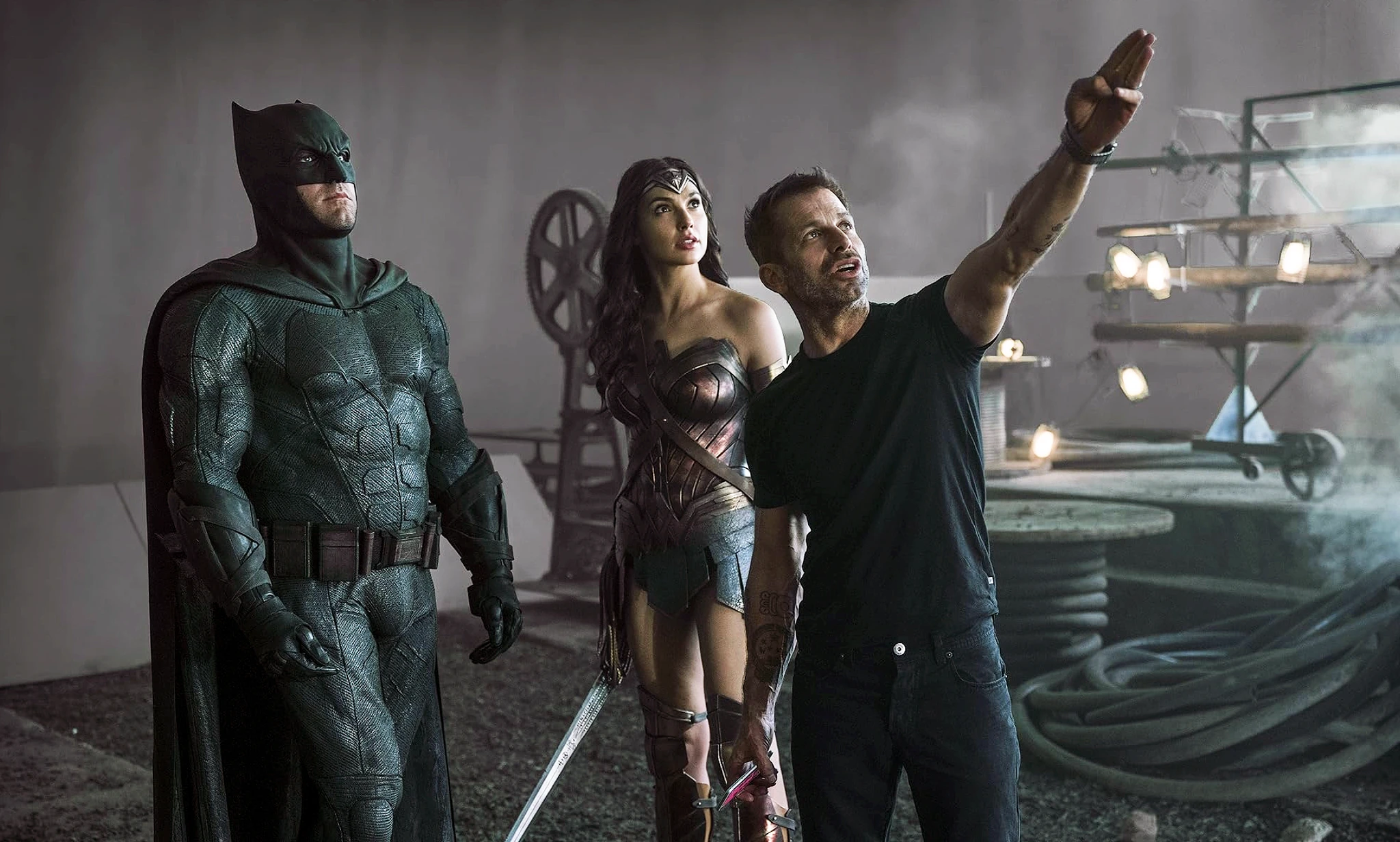 En man i svart t-shirt pekar uppåt och ger instruktioner till två skådespelare i Batman- och Wonder Woman-kostymer, på en filmset med strålkastare och kablar.