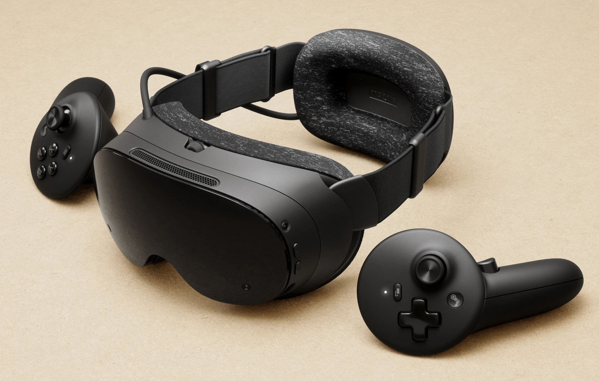 Svart VR-headset Steam Frame med mörkgrått tygklätt huvudband, omgivet av två svarta handkontroller med spak, knappar och styrkors, mot beige bakgrund.